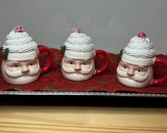 Puede incluir: Tres tazas de cerámica de Papá Noel con tapas en forma de crema batida y asas rojas. Cada taza presenta una cara de Papá Noel con barba blanca y una baya roja encima de la crema batida. Las tazas están sobre una bandeja roja con estampado floral.
