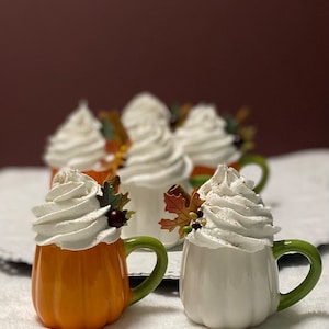 May include: A set of six ceramic pumpkin mugs with white frosting and autumnal decorations. The mugs are arranged in a row on a white surface.