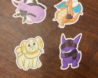 May include: Four colorful cartoon stickers featuring different Pokemon characters. The stickers are on a brown wooden surface. The characters are a purple and black Espeon and Umbreon, a purple and black Gengar, a yellow and white  Jigglypuff, and an orange and blue Charizard.