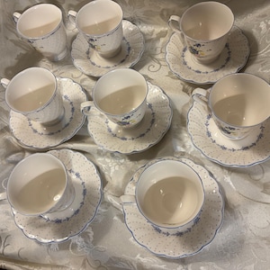 May include: A set of seven cream-coloured ceramic teacups and matching saucers, each featuring a delicate blue polka dot pattern and a floral motif in blue and yellow. The saucers have scalloped edges with a blue trim, and the cups have handles.