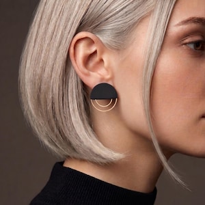 Könnte beinhalten: Ein Ohrstecker mit einem mattschwarzen Halbkreis-Element, an dem zwei dünne, goldfarbene, konzentrische halbkreisförmige Bügel hängen. Der Ohrring wird am Ohrläppchen getragen und zeigt ein geometrisches Design mit kontrastierenden Farben.