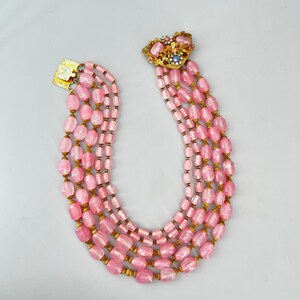 Puede incluir: Un collar de cuentas de vidrio rosa de m&uacute;ltiples hilos con un cierre dorado y un broche floral decorativo. Las cuentas son de forma ovalada y tienen un acabado ligeramente iridiscente.