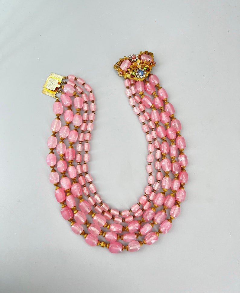 Puede incluir: Un collar de cuentas de vidrio rosa de m&uacute;ltiples hilos con un cierre dorado y un broche floral decorativo. Las cuentas son de forma ovalada y tienen un acabado ligeramente iridiscente.