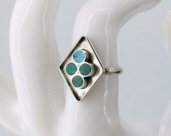 May include: A silver ring with a diamond-shaped face. The face features a turquoise circle and three green circles. The ring is displayed on a white surface.