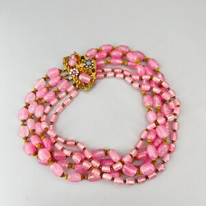 Puede incluir: Un collar de cuentas de vidrio rosa de m&uacute;ltiples hilos con un cierre dorado con un dise&ntilde;o floral con piedras de colores.