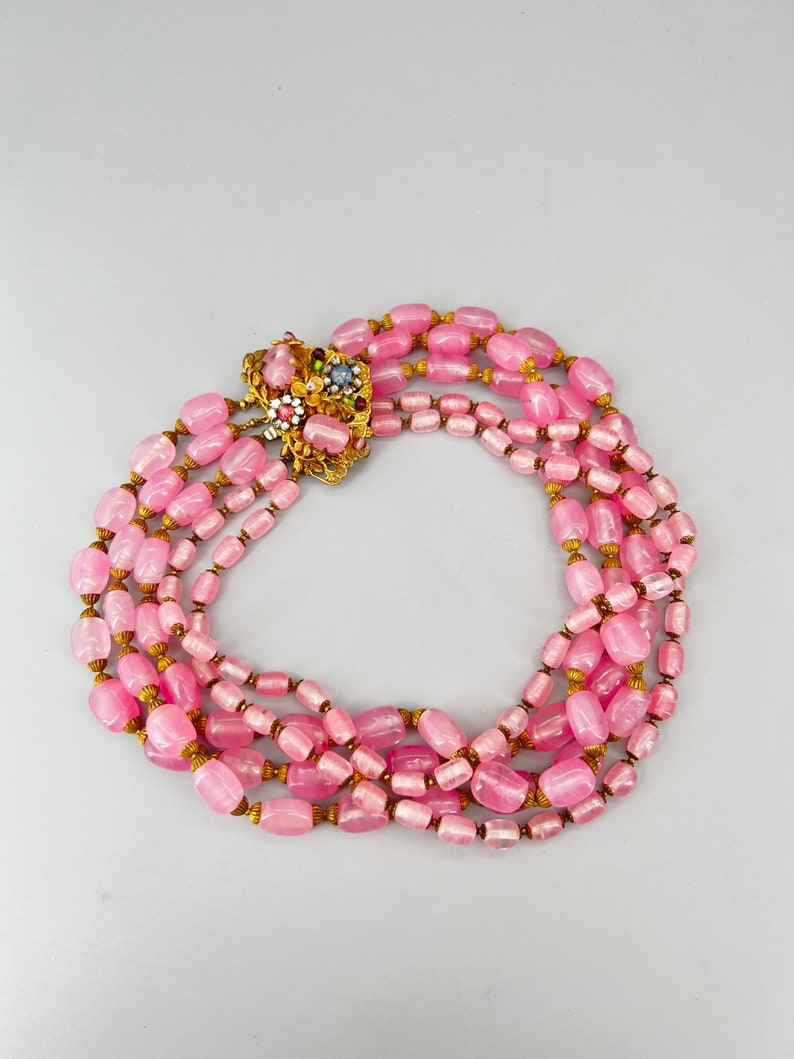 Puede incluir: Un collar de cuentas de vidrio rosa de m&uacute;ltiples hilos con un cierre dorado con un dise&ntilde;o floral con piedras de colores.