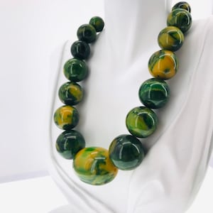 Op de afbeelding: Een ketting van groene en gele gemarmerde plastic kralen. De kralen zijn groot en rond, en de ketting is geregen op een eenvoudig touwtje.