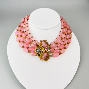 Puede incluir: Un collar choker de cuentas de vidrio rosa de varias hebras con un broche floral dorado. El broche tiene diamantes de imitaci&oacute;n rosas, verdes y azules.