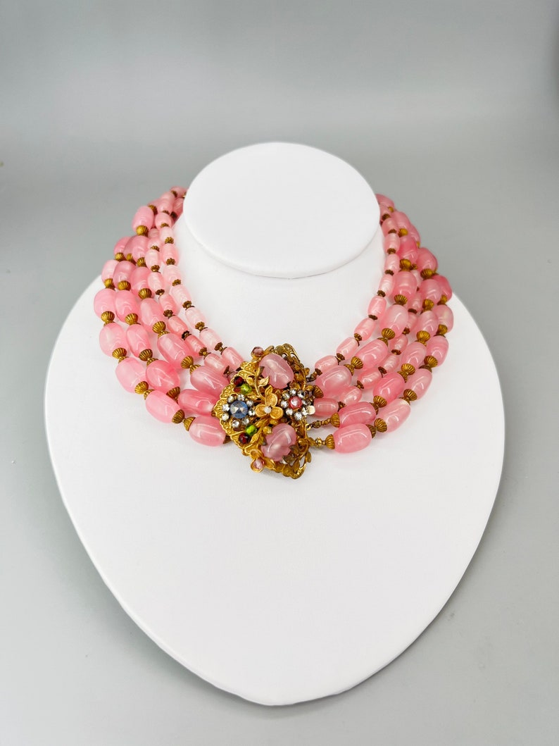 Puede incluir: Un collar choker de cuentas de vidrio rosa de varias hebras con un broche floral dorado. El broche tiene diamantes de imitaci&oacute;n rosas, verdes y azules.