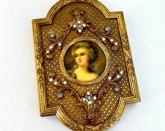 May include: An ornate gold-toned frame with a small portrait of a woman in a white dress. The frame is decorated with rhinestones and blue glass stones.