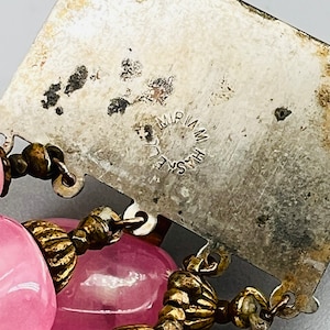 Puede incluir: Un dije de metal plateado con el texto "HASKELL MIRIAM" grabado en &eacute;l. El dije est&aacute; unido a un collar de cuentas de vidrio rosa con detalles de metal dorado.