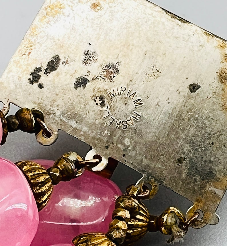 Puede incluir: Un dije de metal plateado con el texto "HASKELL MIRIAM" grabado en &eacute;l. El dije est&aacute; unido a un collar de cuentas de vidrio rosa con detalles de metal dorado.