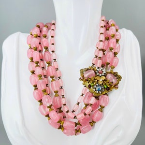 Puede incluir: Un collar de m&uacute;ltiples hilos hecho de cuentas de vidrio rosa con un cierre de metal dorado. El collar presenta un gran colgante de metal dorado adornado con piedras de vidrio de colores.