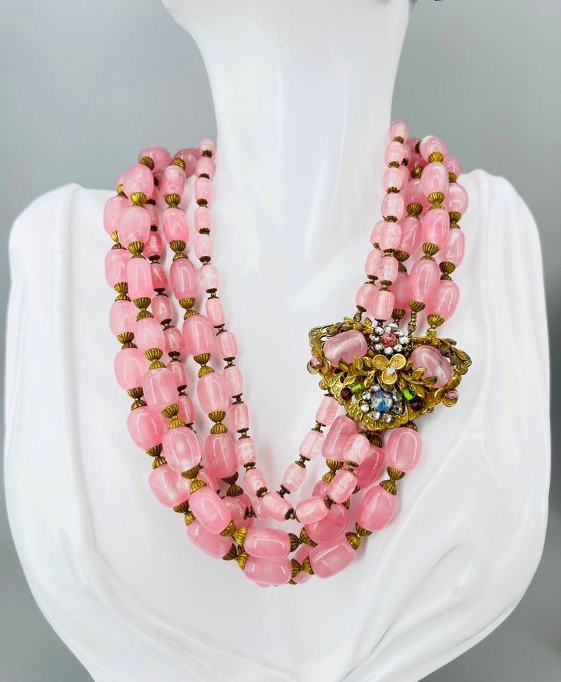 Puede incluir: Un collar de m&uacute;ltiples hilos hecho de cuentas de vidrio rosa con un cierre de metal dorado. El collar presenta un gran colgante de metal dorado adornado con piedras de vidrio de colores.