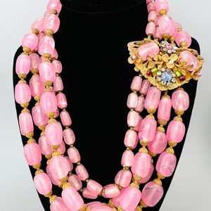 Puede incluir: Un collar de cuentas de vidrio rosa de m&uacute;ltiples hilos con detalles dorados. El collar presenta un broche dorado grande y adornado con piedras de colores.