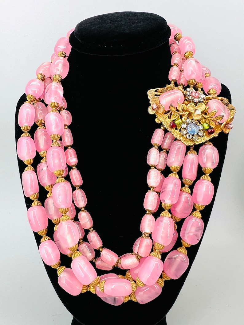 Puede incluir: Un collar de cuentas de vidrio rosa de m&uacute;ltiples hilos con detalles dorados. El collar presenta un broche dorado grande y adornado con piedras de colores.