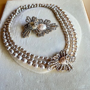 May include: A three-strand pearl necklace with a large, ornate, crystal-encrusted brooch. The brooch features a central pearl and a delicate, floral design.