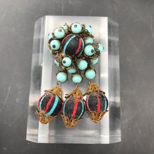 Puede incluir: Un broche vintage con un racimo de cuentas de vidrio azul turquesa y negras, acentuadas con metal dorado. El broche presenta tres adornos colgantes con patrones de rayas negras, rojas y azul turquesa.