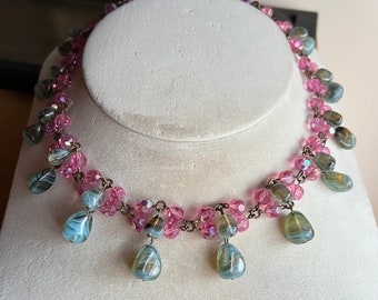 May include: A pink and green glass beaded necklace with teardrop-shaped pendants. The necklace is on a white mannequin bust.