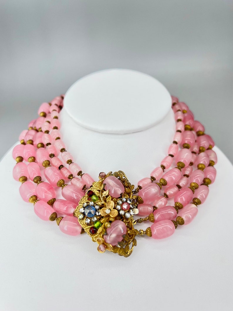 Puede incluir: Un collar de cuentas de vidrio rosa de m&uacute;ltiples hilos con un cierre floral dorado adornado con brillantes de colores.