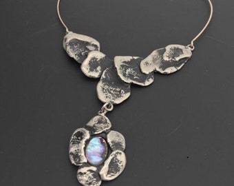 Puede incluir: Un collar de plata con un colgante que presenta un grupo de formas irregulares texturizadas y una piedra iridiscente central.