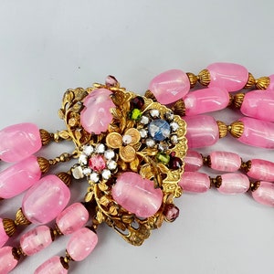 Puede incluir: Un collar de m&uacute;ltiples hilos con cuentas de vidrio rosa y un cierre adornado en oro con piedras de colores.