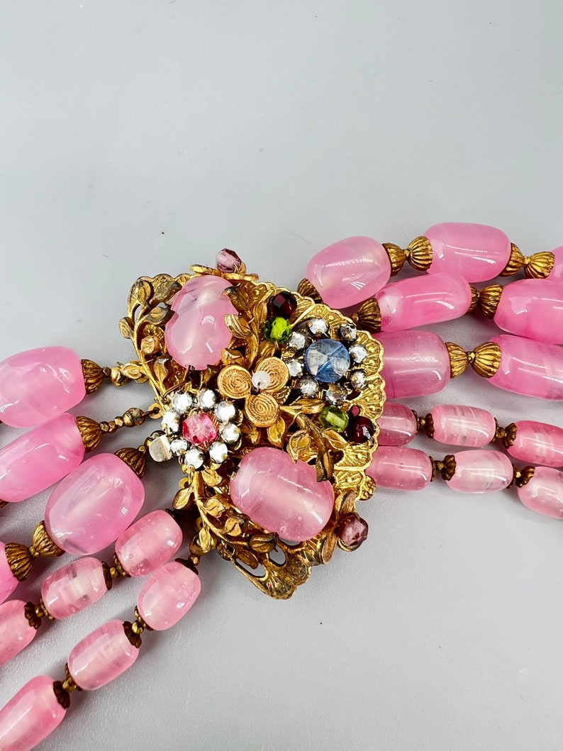 Puede incluir: Un collar de m&uacute;ltiples hilos con cuentas de vidrio rosa y un cierre adornado en oro con piedras de colores.