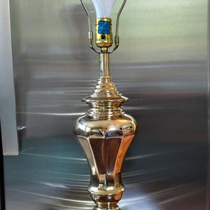 May include: A polished brass table lamp featuring a multi-tiered base and a slender, contoured body. The lamp includes a gold-toned metal socket with a blue label, a harp attachment, and a single white light bulb extending from the top fixture.