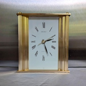 May include: A rectangular Seiko quartz mantel clock featuring a white face with black Roman numerals and thin black hands. The clock is encased in a gold-toned frame with vertical fluted columns on the left and right sides, set on a flat base.