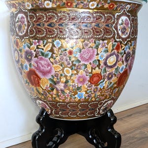 May include: A large, round ceramic fishbowl planter featuring a gold-toned background with a dense, colorful floral pattern of pink, red, and purple blossoms. The rim has a decorative border, and the bowl rests on a black, carved wooden stand.