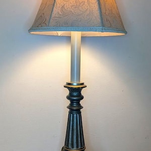 May include: A table lamp with a dark metallic, fluted base featuring ornate carved details and a round, stepped pedestal. The lamp is topped with a conical, off-white fabric shade that displays a subtle, repeating floral pattern and emits a warm glow.