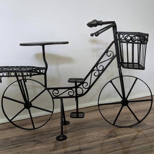 May include: A black metal decorative bicycle stand featuring a front wire basket, a flat rear rack, a small seat, and thin spoked wheels. The frame is constructed with curved metal scrollwork details and includes a pedal and a support stand.