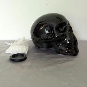 May include: A glossy black ceramic skull sculpture with a hollow interior, featuring a small circular opening on the side. Beside the skull sits a small, clear drawstring bag filled with tiny translucent beads and a separate black circular lid.