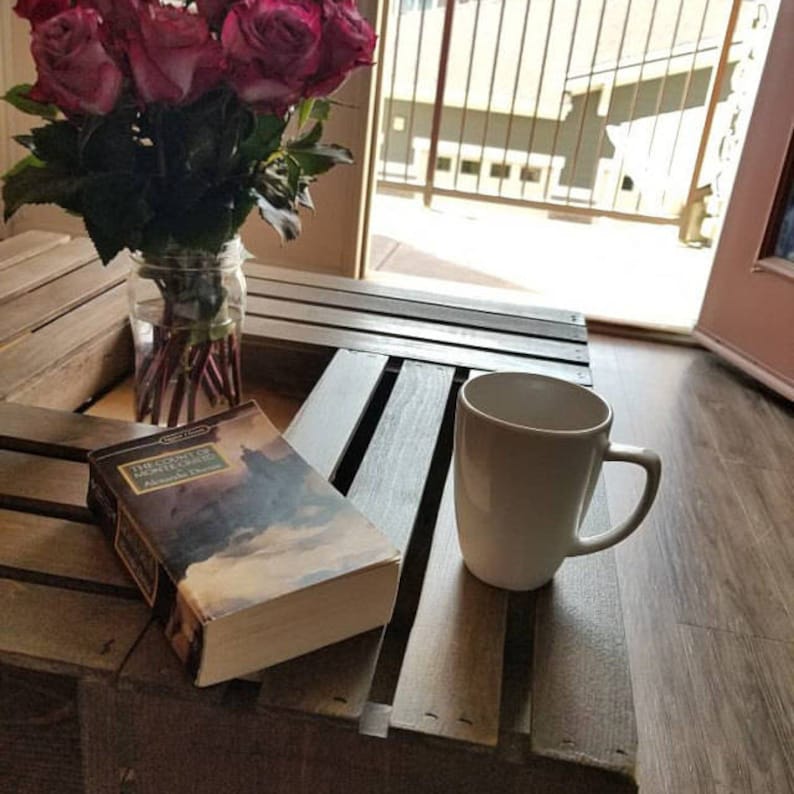 Op de afbeelding: Een salontafel van houten kratten met een witte mok, een boek getiteld "The Lord of the Rings" en een boeket roze rozen in een glazen vaas.