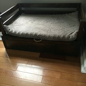 May include: A dark brown wooden dog bed with a gray faux fur mattress and a storage drawer underneath.