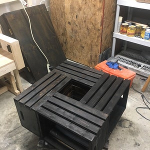 Op de afbeelding: Een zwarte houten salontafel gemaakt van vier gestapelde kratten. De kratten zijn in een vierkante vorm gerangschikt met de bovenste krat open in het midden. De tafel staat op een betonnen vloer.