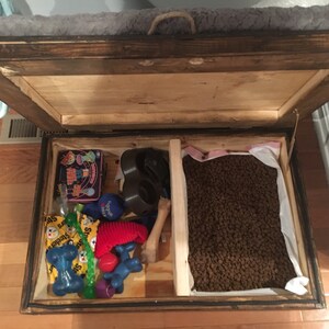 May include: Open wooden storage box with a divider. One side contains dog toys, including a bone, a red ball, and a blue rope toy. The other side contains a bag of dog food.