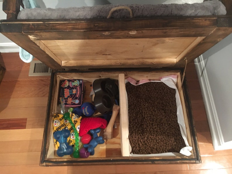 May include: Open wooden storage box with a divider. One side contains dog toys, including a bone, a red ball, and a blue rope toy. The other side contains a bag of dog food.