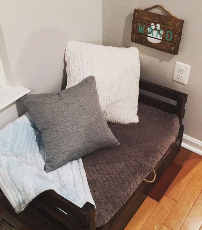 May include: A wooden dog bed with a brown and gray blanket, a gray pillow, and a white pillow. The bed has a drawer and a sign that says "WARD" with a paw print.