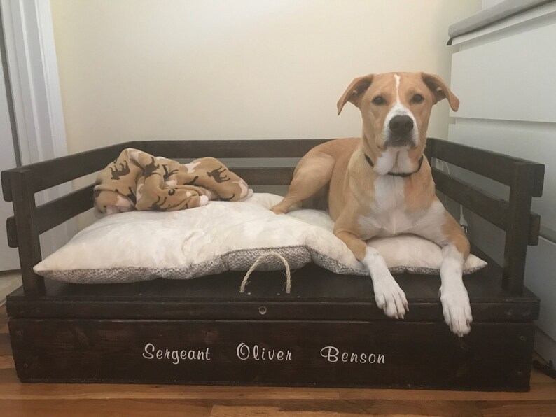 May include: A dark brown wooden dog bed with a white plush cushion and a tan and brown blanket. A light brown dog with white markings is lying on the bed. The bed has a storage drawer with the names "Sergeant Oliver Benson" written on it.