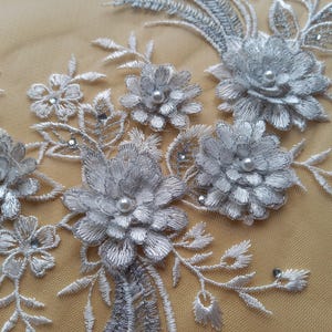 Puede incluir: Primer plano de encaje floral plateado con flores y hojas bordadas. Cada flor tiene un centro de perla. El encaje está sobre un fondo beige, creando un diseño delicado y elegante.