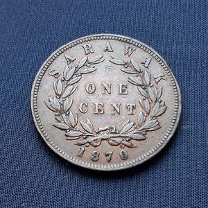 Könnte beinhalten: Silberne Sarawak One Cent Münze von 1870. Die Münze zeigt das Wort "SARAWAK" oben, einen Kranz und die Worte "ONE CENT" in der Mitte. Das Jahr "1870" befindet sich unten. Die Münze liegt auf einer blauen Oberfläche.