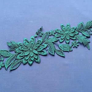 May include: Emerald green floral lace applique. The applique features detailed flowers and leaves, creating a decorative design. The lace is set against a blue background, highlighting the intricate details of the embroidery.