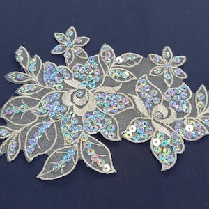 May include: A floral applique with a delicate design. The applique features white embroidered flowers and leaves, embellished with iridescent sequins. The sequins catch the light, creating a shimmering effect. The applique is set against a dark blue background.
