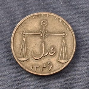 May include: A round bronze-colored coin featuring the text QUARTER ANNA at the top. The center displays an embossed image of a balance scale with Arabic script positioned between the two pans. The date 1246 is inscribed in Arabic numerals at the bottom.