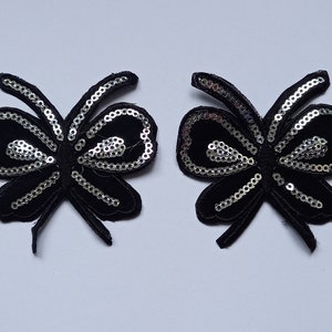 May include: Two black velvet butterfly appliques with silver sequins outlining the wings. The appliques are symmetrical and have a soft, textured appearance. These decorative embellishments are suitable for various craft projects.