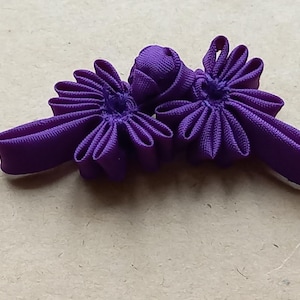 Könnte beinhalten: Zwei lila Stoffknöpfe in Blumenform. Jeder Knopf hat einen zentralen Knoten und strahlenförmige Blütenblätter. Die Knöpfe sind aus einem glatten, einfarbigen Stoff gefertigt. Die Knöpfe befinden sich auf einem neutralen Hintergrund.
