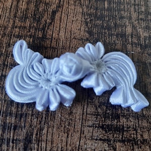 May include: Two light purple satin fabric embellishments. Each has a central flower shape and radiating petal-like elements. The embellishments are connected and rest on a wooden surface.