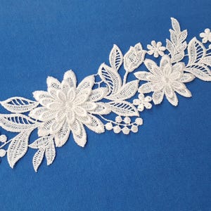 Könnte beinhalten: Weiße florale Spitzenapplikation mit detaillierten Blumen- und Blattmotiven. Die Spitze ist in einem geschwungenen Muster auf blauem Hintergrund angeordnet. Diese dekorative Spitze eignet sich für verschiedene Bastelprojekte.