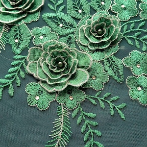 May include: Close-up of green embroidered floral lace. The design features detailed roses, leaves, and small embellishments. The embroidery is outlined with silver thread, creating a textured and dimensional effect. The lace is set against a dark green fabric.